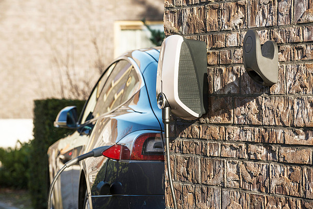 Tankstelle für ein Elektroauto an einer Hauswand, Elektroauto mit Ladekabel im Hintergrund